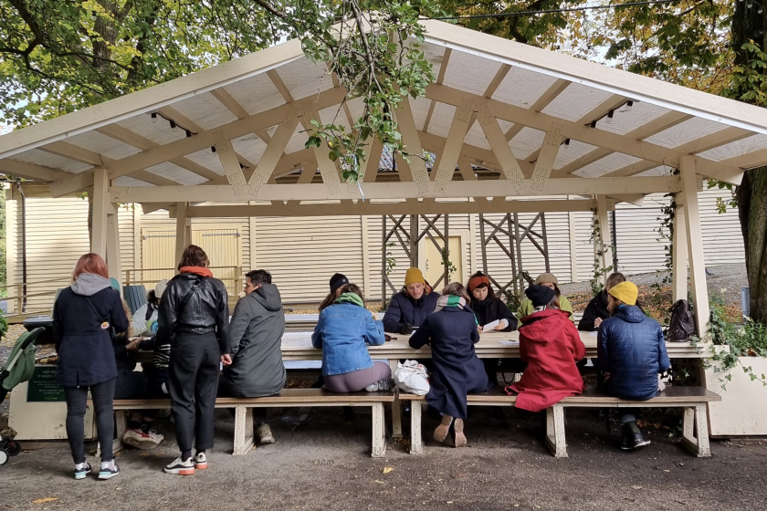 Group of people sitting at a bench under a shelter, outside in a park 