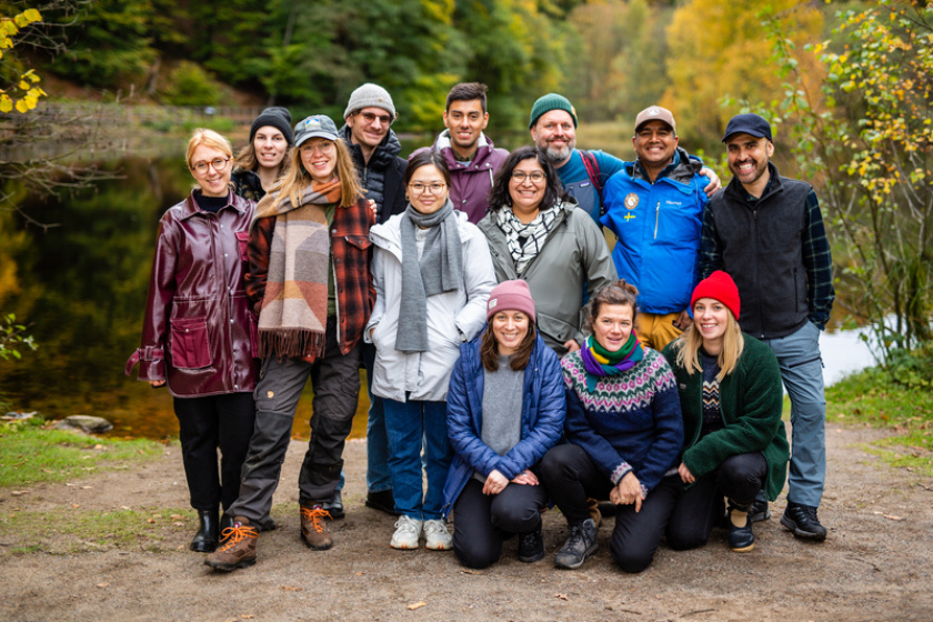 Thirteen people posing in front of a lake in fall. 