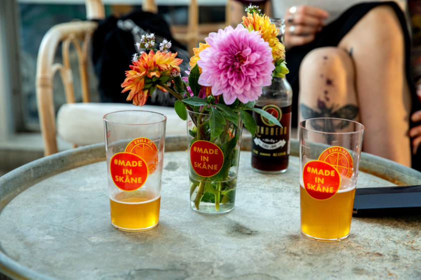 Ölglas med texten Made in Skåne, på ett bord med en vas och blommor
